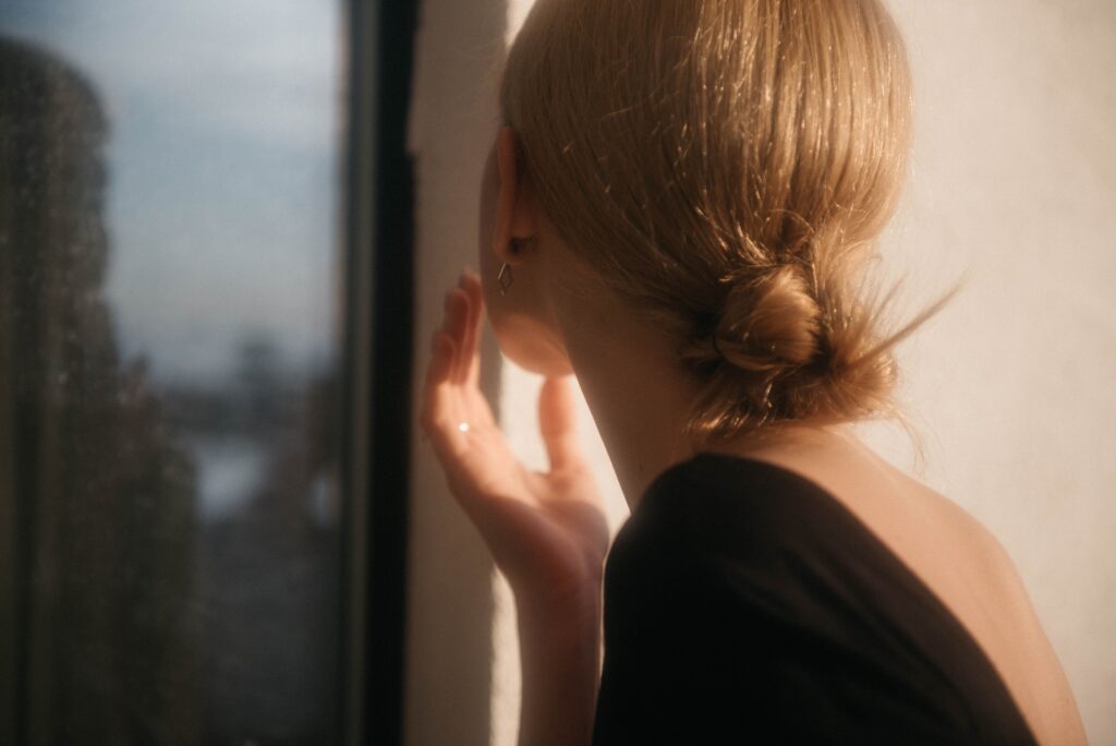 How to Get a Guy to Like You: 15 Ways (Without Looking Clingy) Back view of a woman looking out a sunlit window, reflecting on how to get a guy to like you.