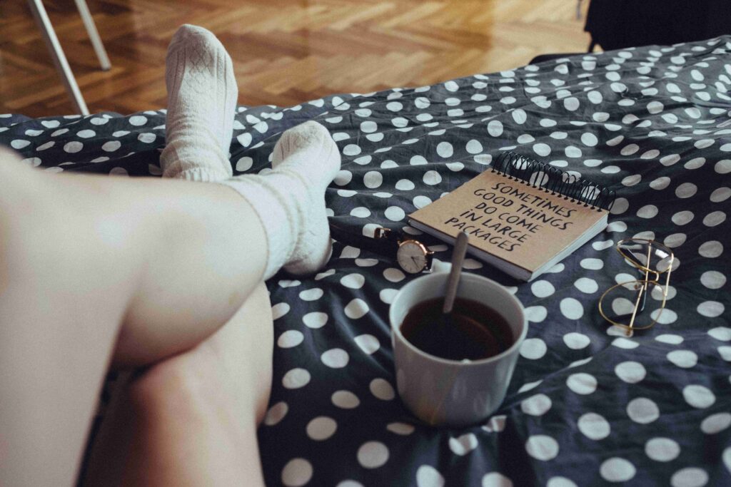115 Powerful Monday Morning Quotes to Beat the Blues A cozy bedroom view of legs in wool socks on a polka dot blanket with coffee, reading positive Monday morning quotes.