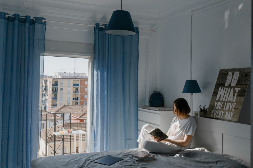 135 Wednesday Motivational Quotes for Midweek Energy A woman reading a book on her bed with blue curtains, gaining fresh energy from Wednesday motivational quotes.