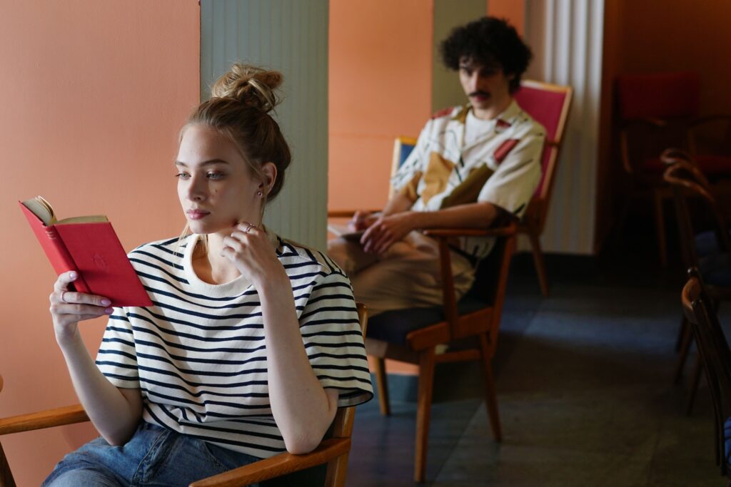 “Why Does My Girlfriend Hate Me?” 9 Reasons for Her Sudden Distance A distant young woman reading a book while her partner sits in the background, thinking why does my girlfriend hate me.