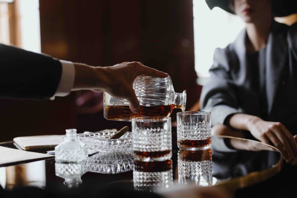 A hand pouring whiskey from a decanter into glasses at a dark table, highlighting a sovereign woman lifestyle.