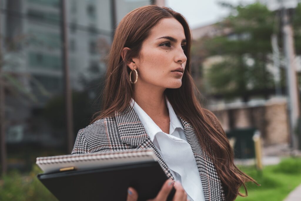 Alpha Female Meaning: 17 Signs & How She Loves A confident woman in a plaid blazer holding a notebook outdoors, capturing the alpha female meaning.