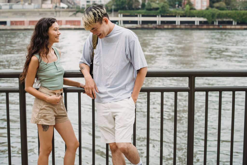 A young guy and girl laughing together next to a waterfront railing while finding a romantic partner.