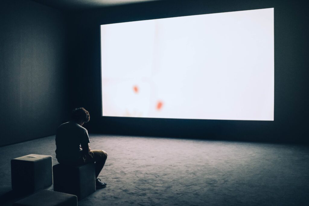 A person sitting alone in a dark room facing a giant blank projection screen, tracing reasons for being single.
