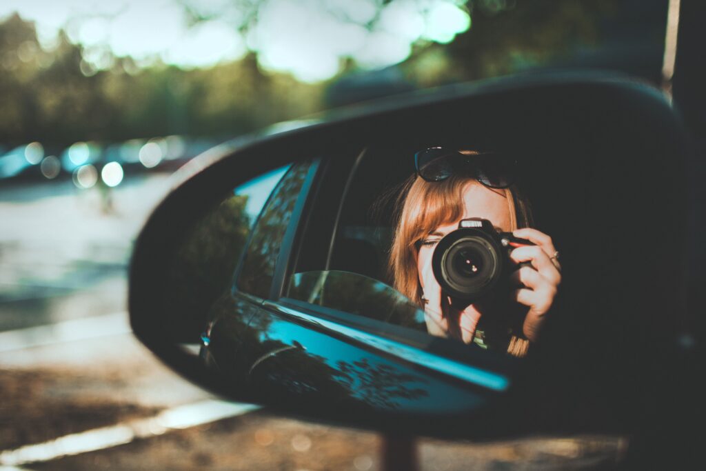 Reflection in a car side mirror of a woman holding a camera to her face, focuses on manifesting true connection.