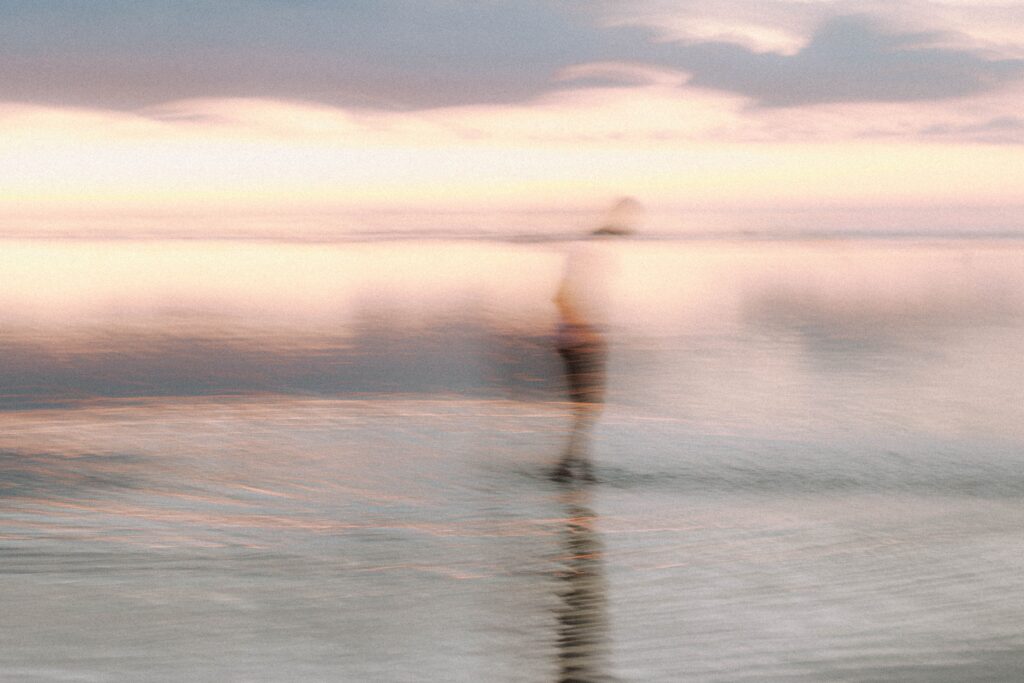 A heavily blurred and motion-streaked figure walking on a calm sea during a hazy dusk, a vision of late loved ones well.