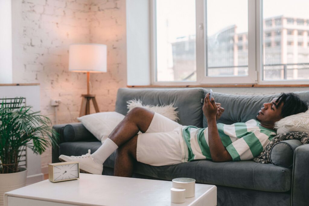A young man lying on a gray couch checking his phone for signs of interest and dating tips for men.