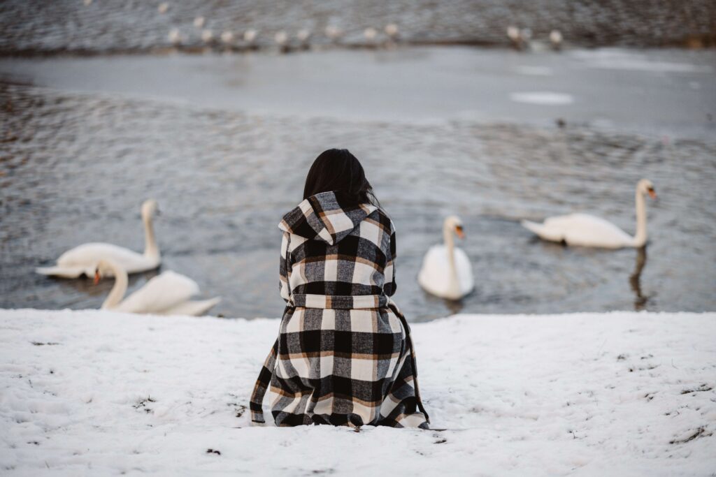 Why Am I Single? 7 Hidden Mental Blocks And How to Fix Them Back view of a lonely woman sitting on snow looking at swans, reflecting on why am I single.