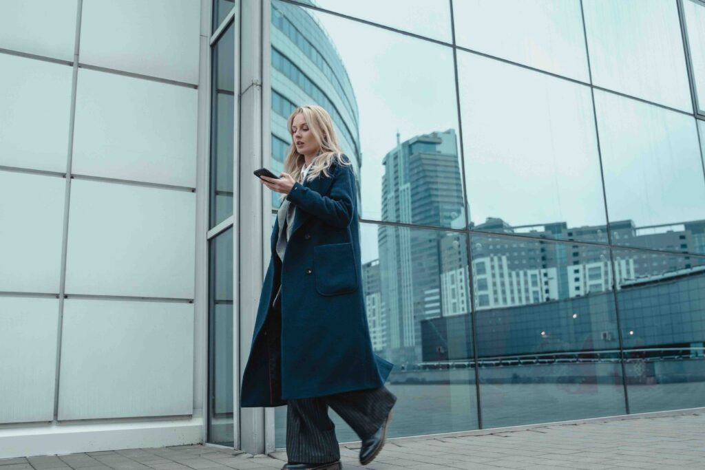 A confident woman in a blue coat walking outside a modern glass building, showing independent woman characteristics.