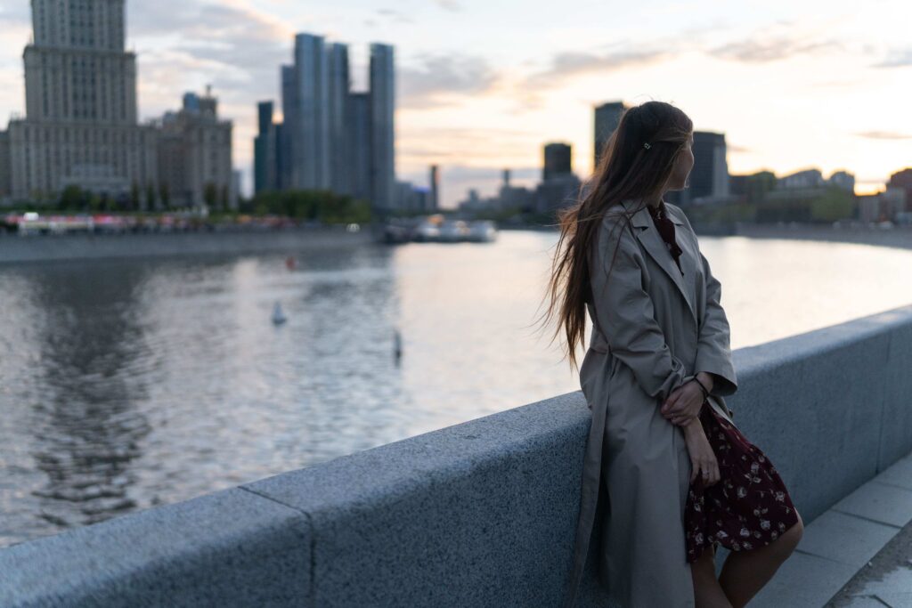 Side view of a girl in a coat looking out at the river and city skyline, dreaming of discovering lasting romance.