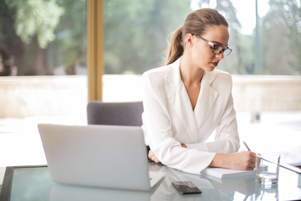 A focused woman in a white blazer writing in a notebook near a laptop, showing an assertive woman definition.