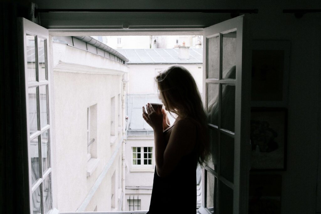 Silhouette of a woman drinking coffee by an open balcony window, processing hidden psychological relationship blocks.