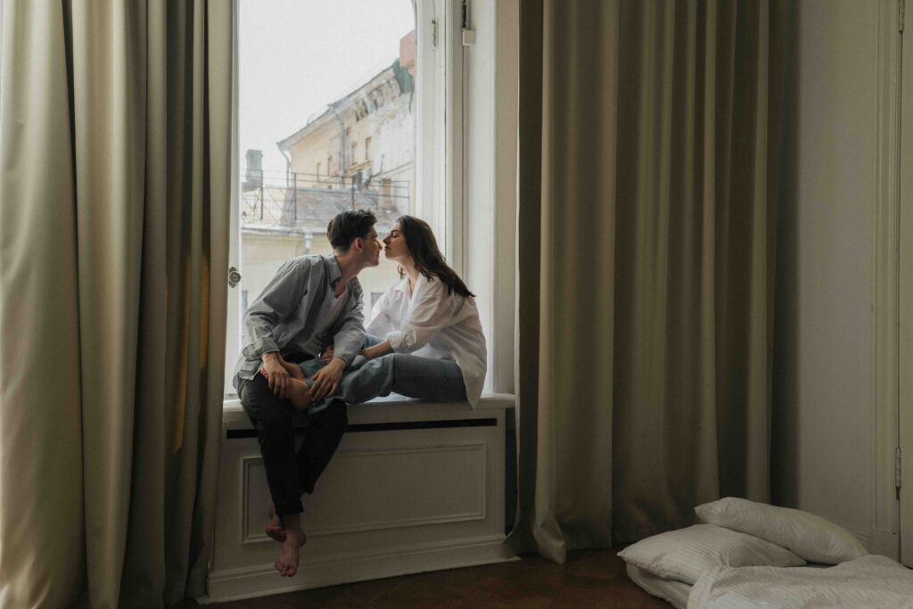 A couple sitting on a window sill getting close, testing their connection and initial romantic attraction.