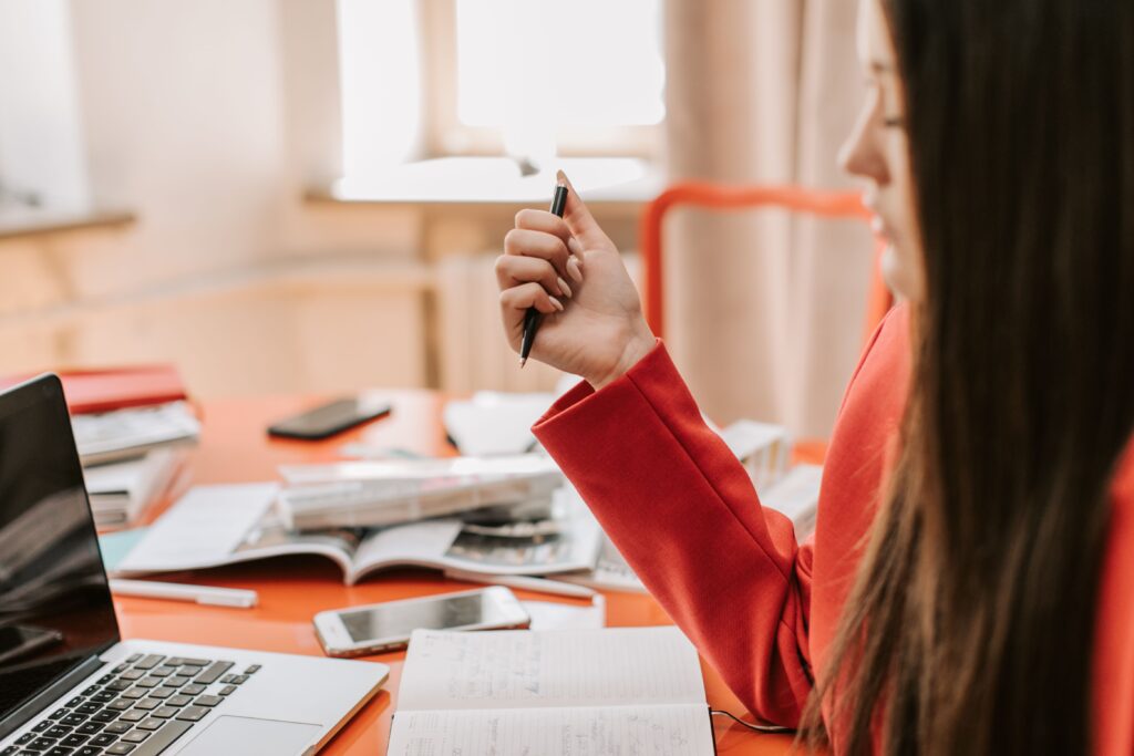 Side view of a woman with a pen studying at an orange desk with a laptop, reflecting self reliant female traits.