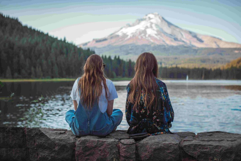 Kindred Spirits Meaning: 7 Signs & How It Differs From Soulmates Two friends sitting near a lake facing mountains reflecting kindred spirits meaning and mental alignment.