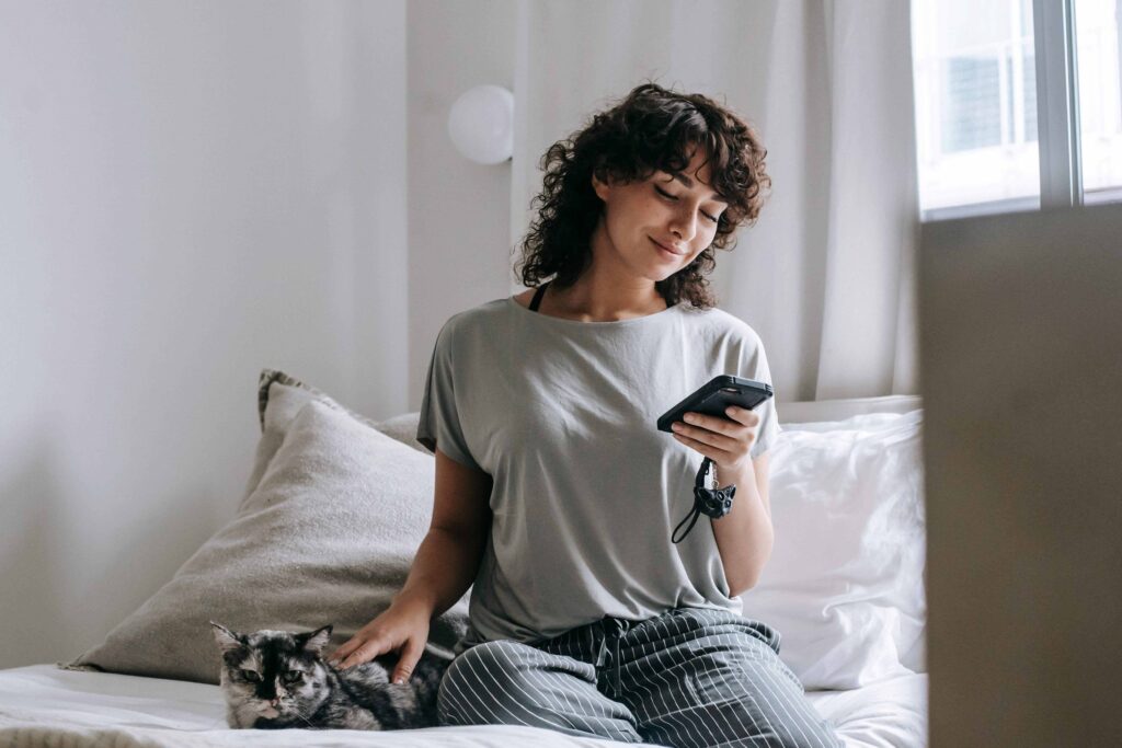 A happy woman sitting on bed with her cat receiving cute morning love lines for girlfriend on the phone. 