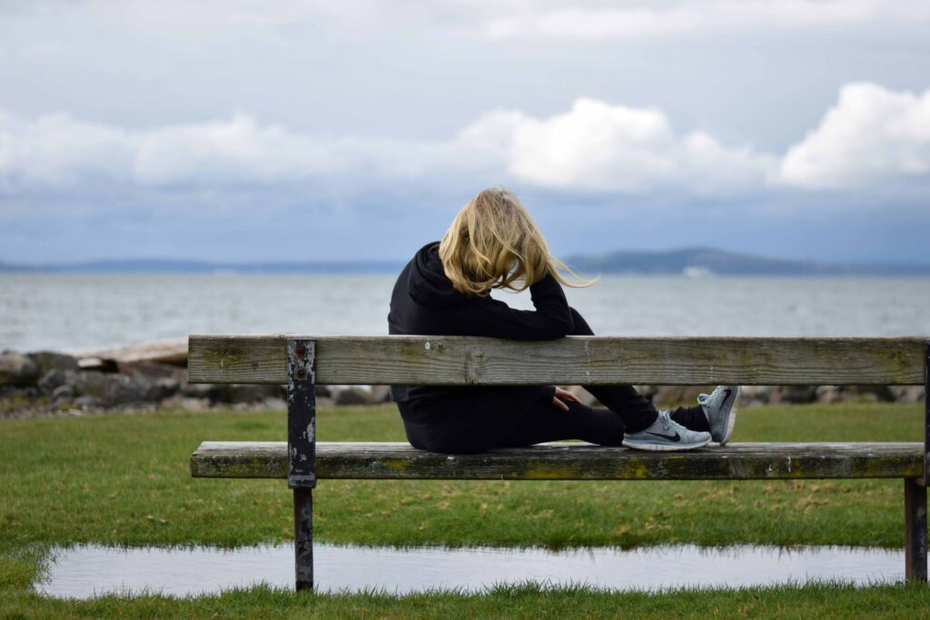 A woman sitting alone on a park bench by the water reflecting identity crisis meaning.