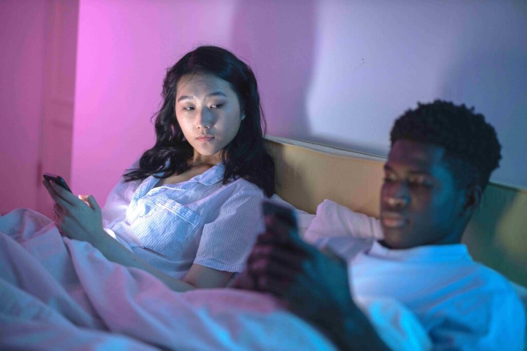 What Is Micro-Cheating? 11 Sneaky Behaviors Ruining Relationships A couple in bed looking at their phones reflecting what is micro-cheating.