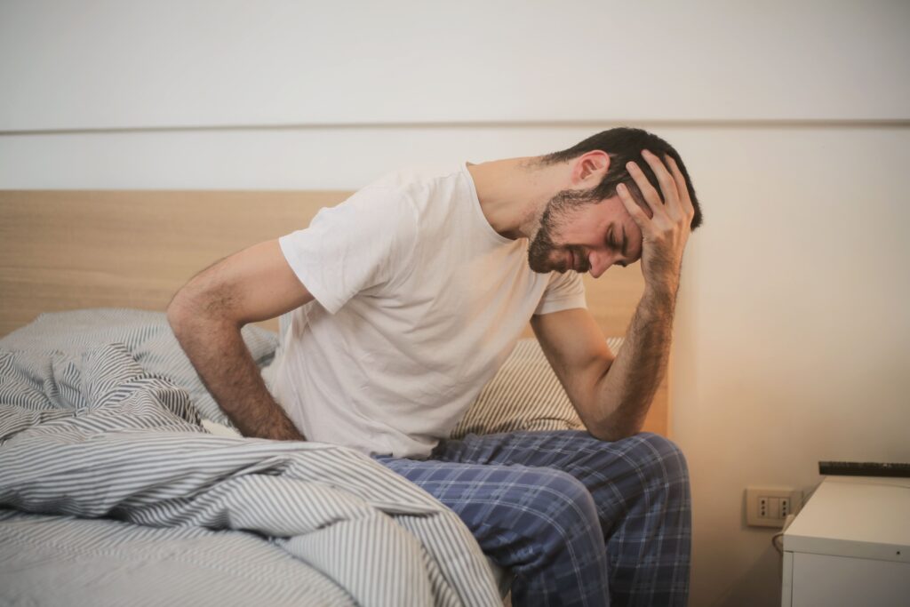 A stressed man holding head while sitting on edge of bed analyzing rem sleep cycles.