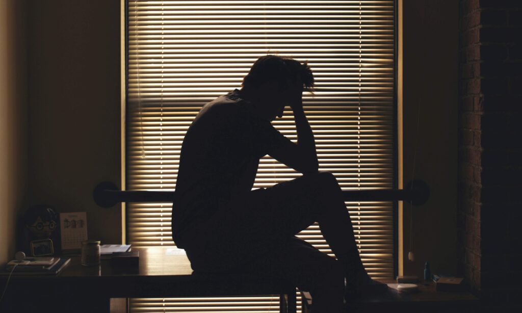 A silhouette of a distressed man sitting by window blinds coping with acute loneliness.